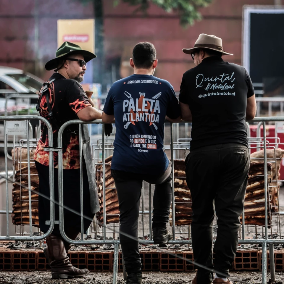 Assadores observam as costelas no fogo de chão, um deles com a camiseta Assador Celebridade do Paleta Atlântida