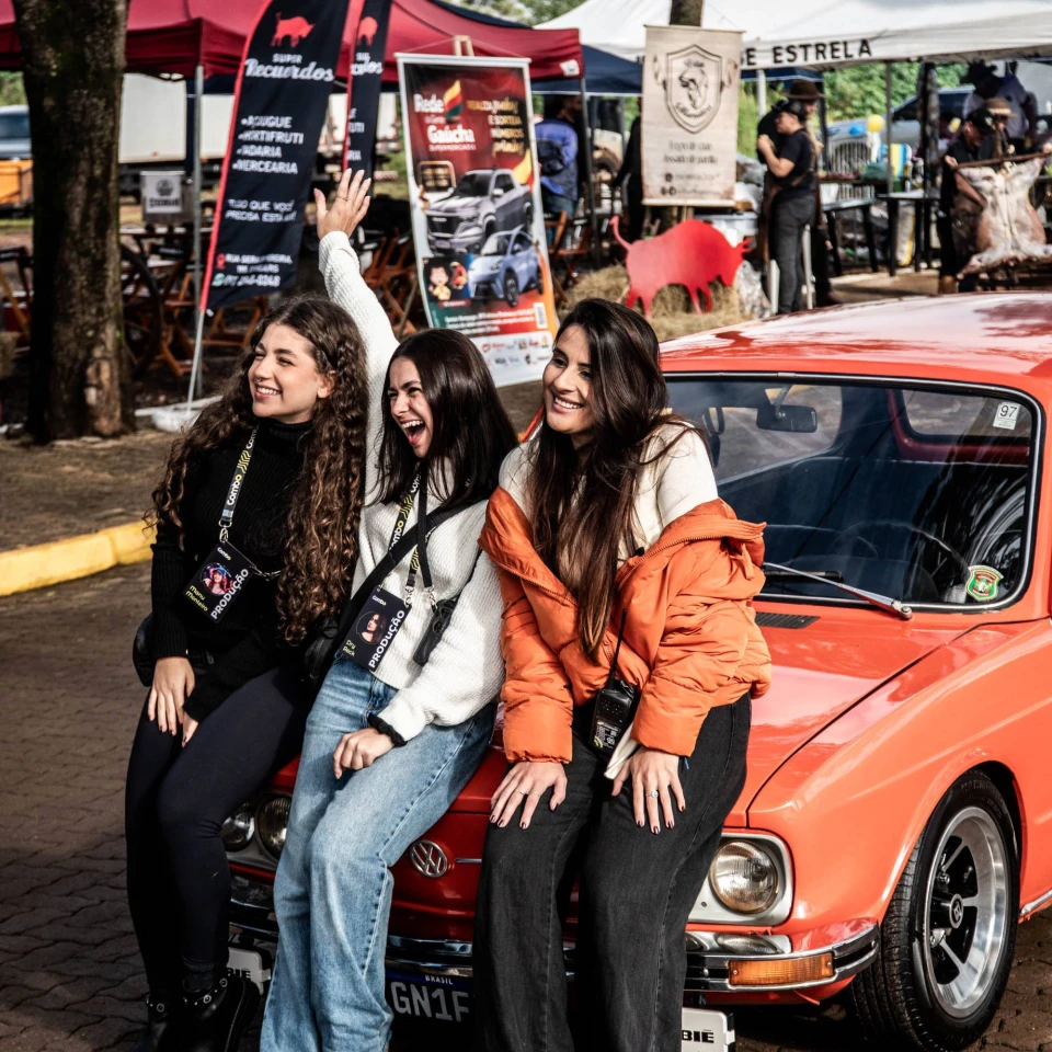 Equipe de produção do Paleta Atlântida comemora sentada no capô de um carro antigo laranja