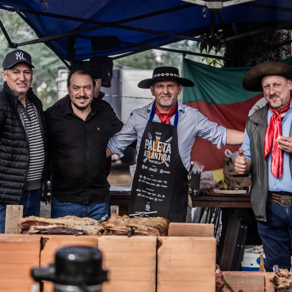 Assadores pilchados posam diante das carnes no fogo de chão, com a bandeira do Rio Grande do Sul ao fundo